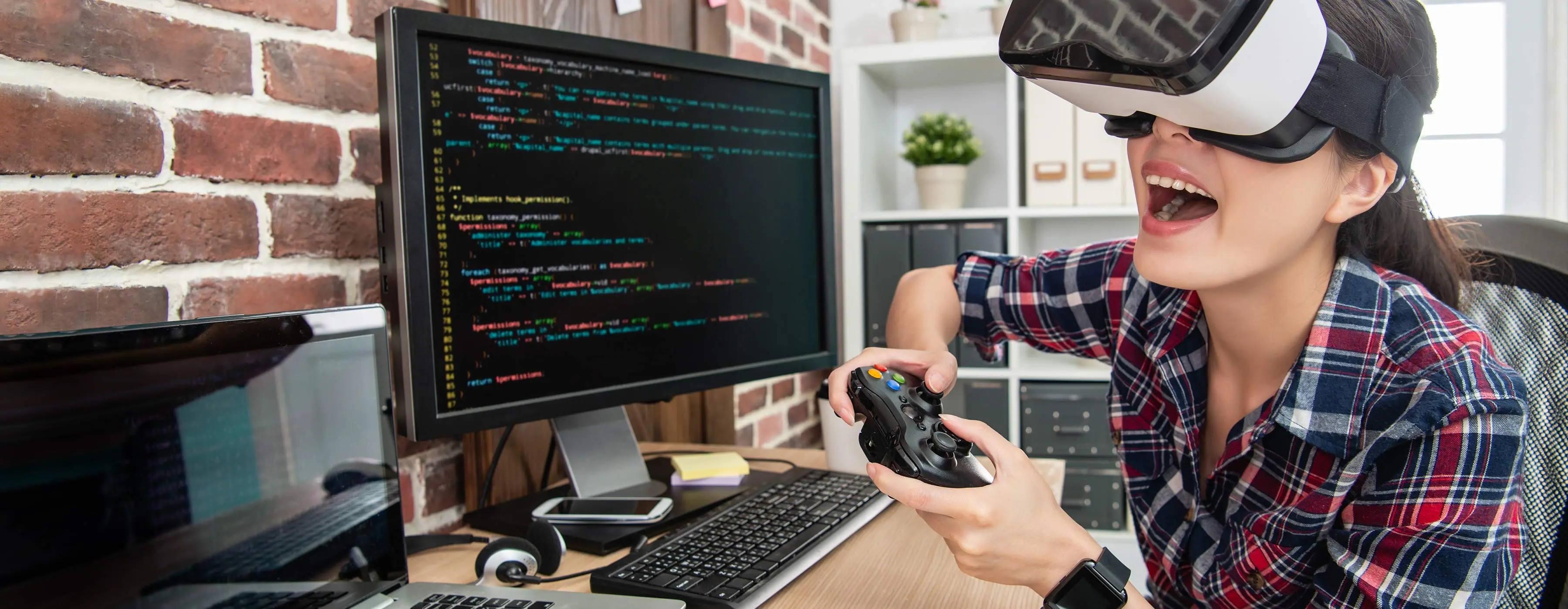 Foto de uma jovem em escritório com óculos de realidade virtual 3D e controle sem fio de videogame em frente a computador e notebook com códigos de programação na tela.