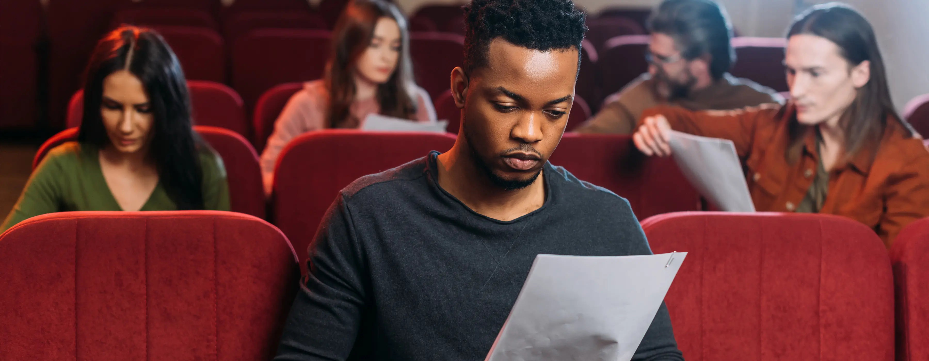 Foto de um homem jovem lendo um papel no auditótio de um teatro, com grupo de pessoas ao fundo