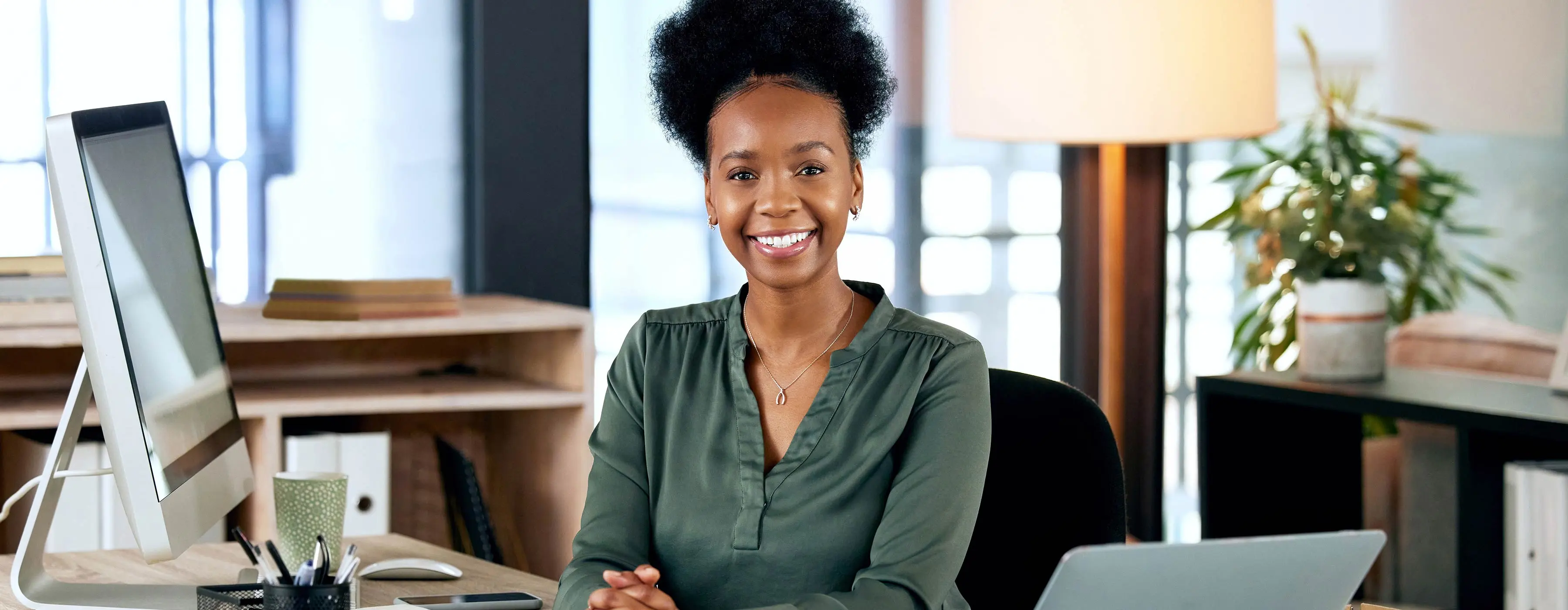 Foto de mulher sorrindo, em um cenário de escritório, com um computador à sua frente.