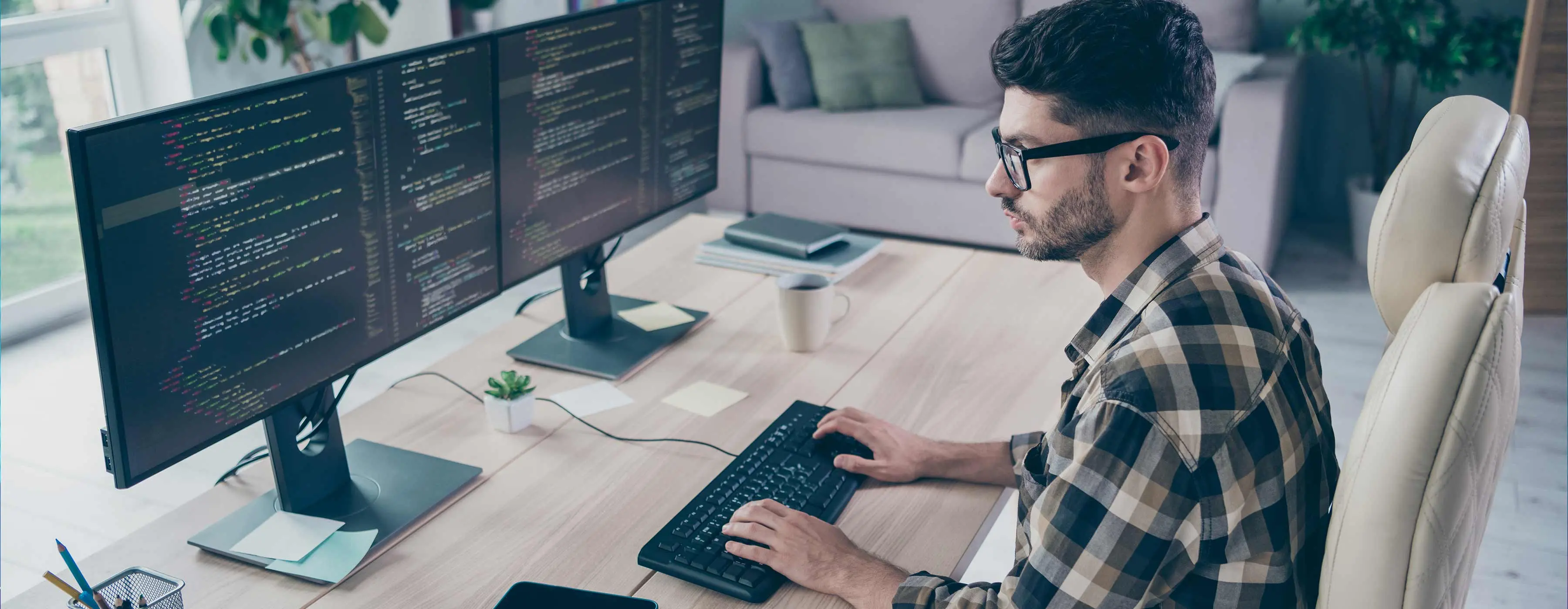 Foto de um homem digitando códigos de programação em duas telas de computador em escritório.