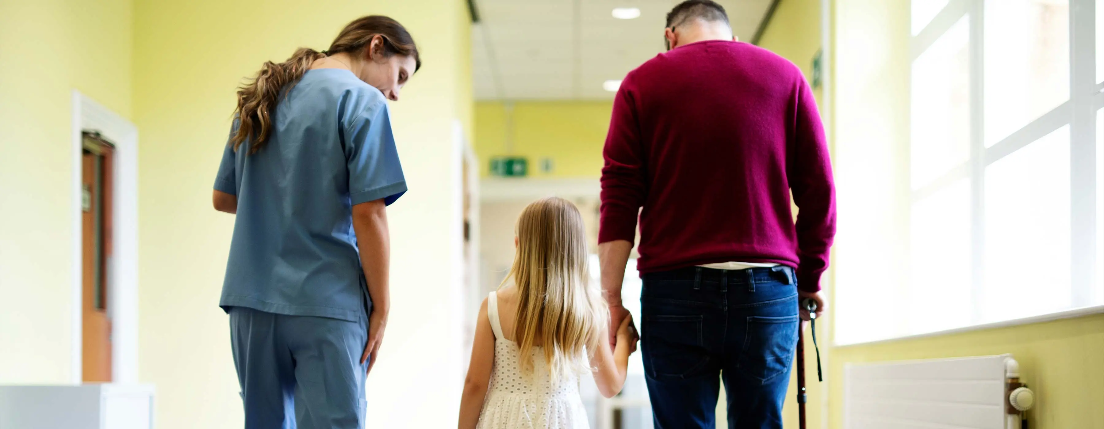 Foto de mulher com uniforme azul convesando e caminhando ao lado de uma menina que está de mãos dadas com homem de muleta em corredor de hospital.