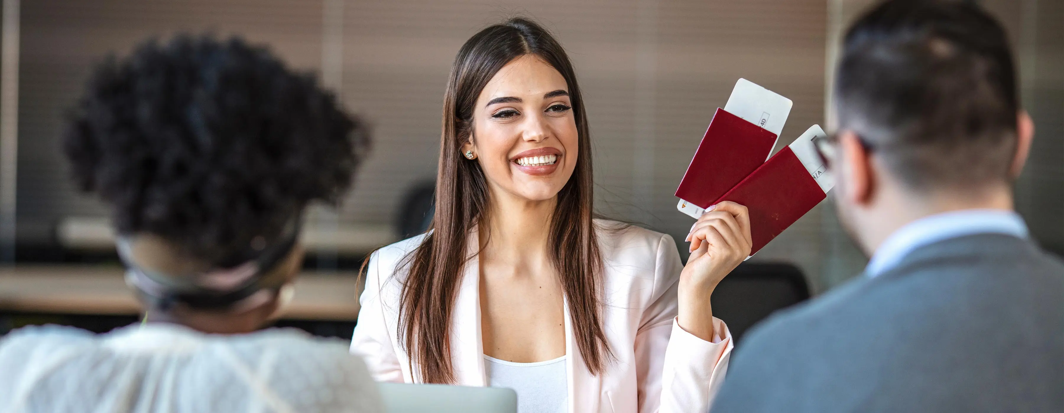 Foto de mulher sorrindo segurando dois passaportes com ticket de passagens mostrando para duas pessoas em escritório.