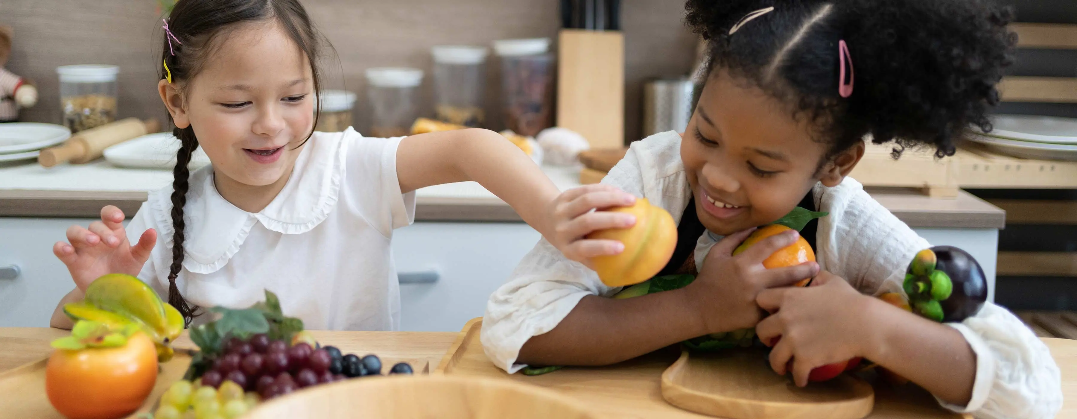Foto de duas crianças brincando com frutas em cima de uma mesa, em um cenário de cozinha ao fundo.
