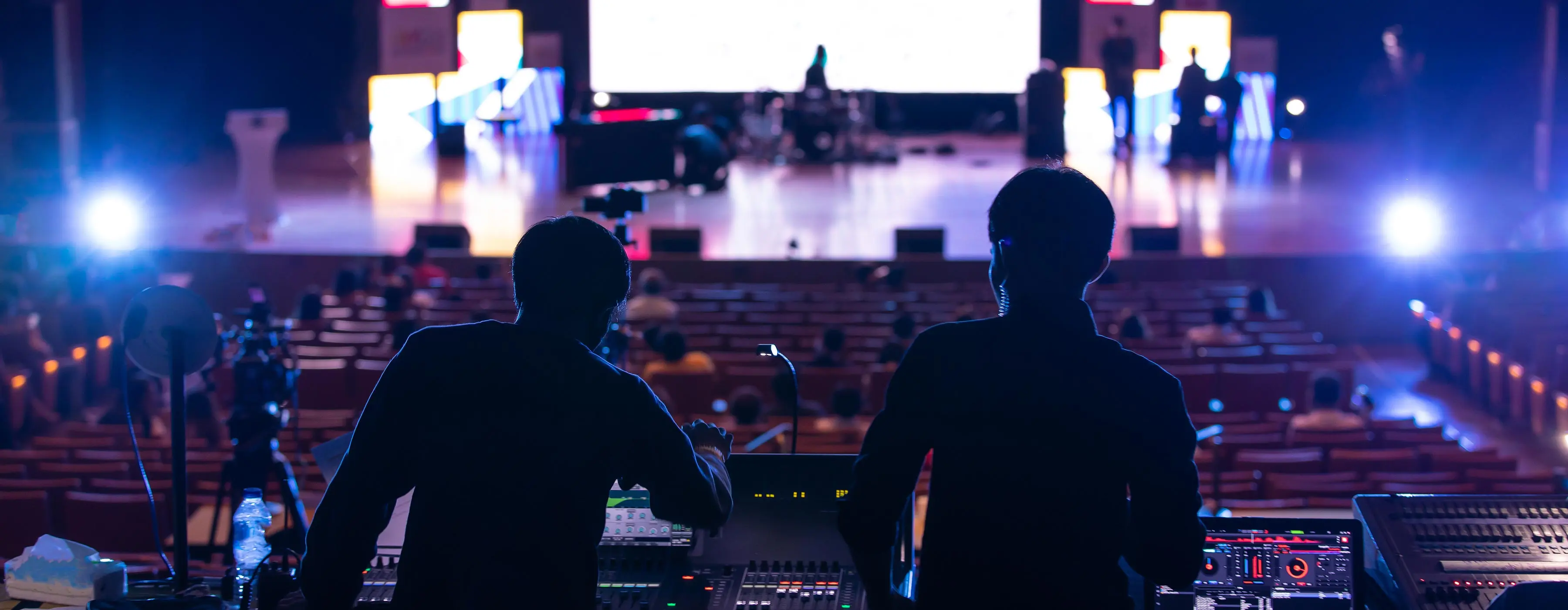 Foto de duas pessoas operando mesa de som ao fundo da plateia de um teatro.