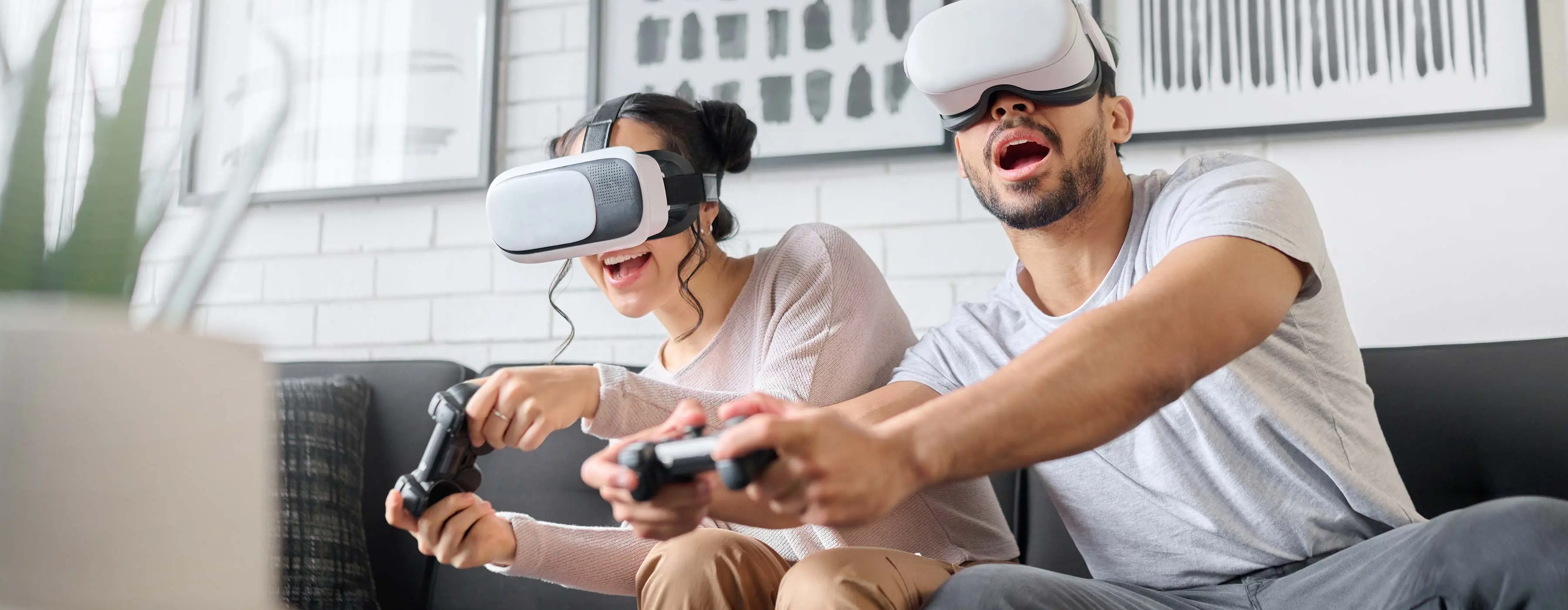 Foto de uma moça e um rapaz sentados no sofá  jogando videogame utilizando óculos de realidade virtual e controle sem fio.
