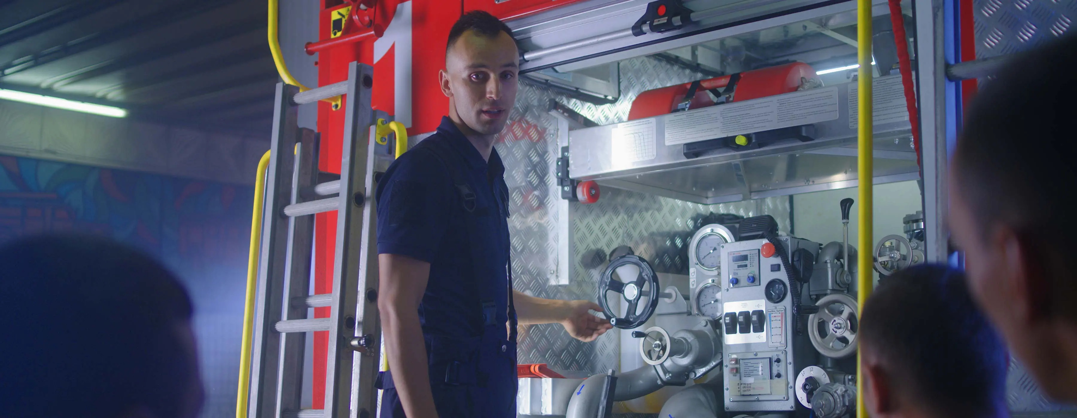 Foto de homem uniformizado mostrando equipamentos de uma viatura de bombeiro para pessoas sentadas.