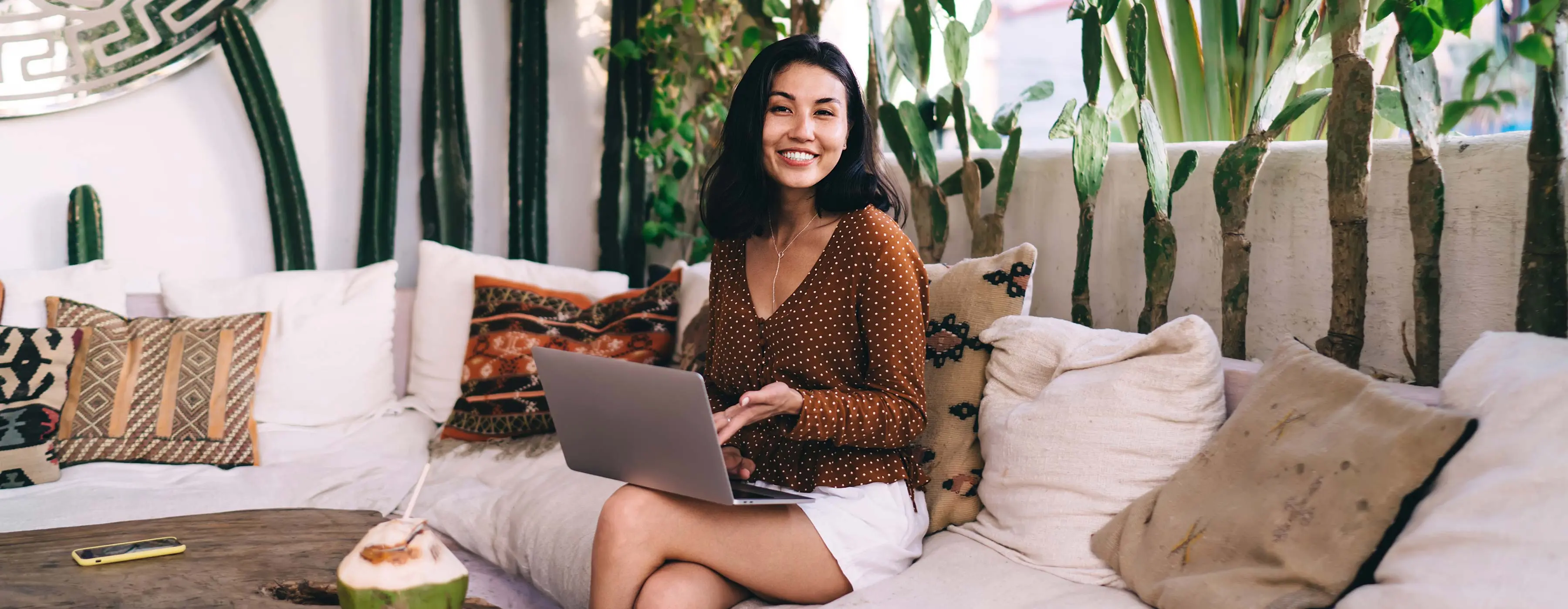 Foto de mulher usando notebook sentada em sofá rodeado por vasos com cactos.