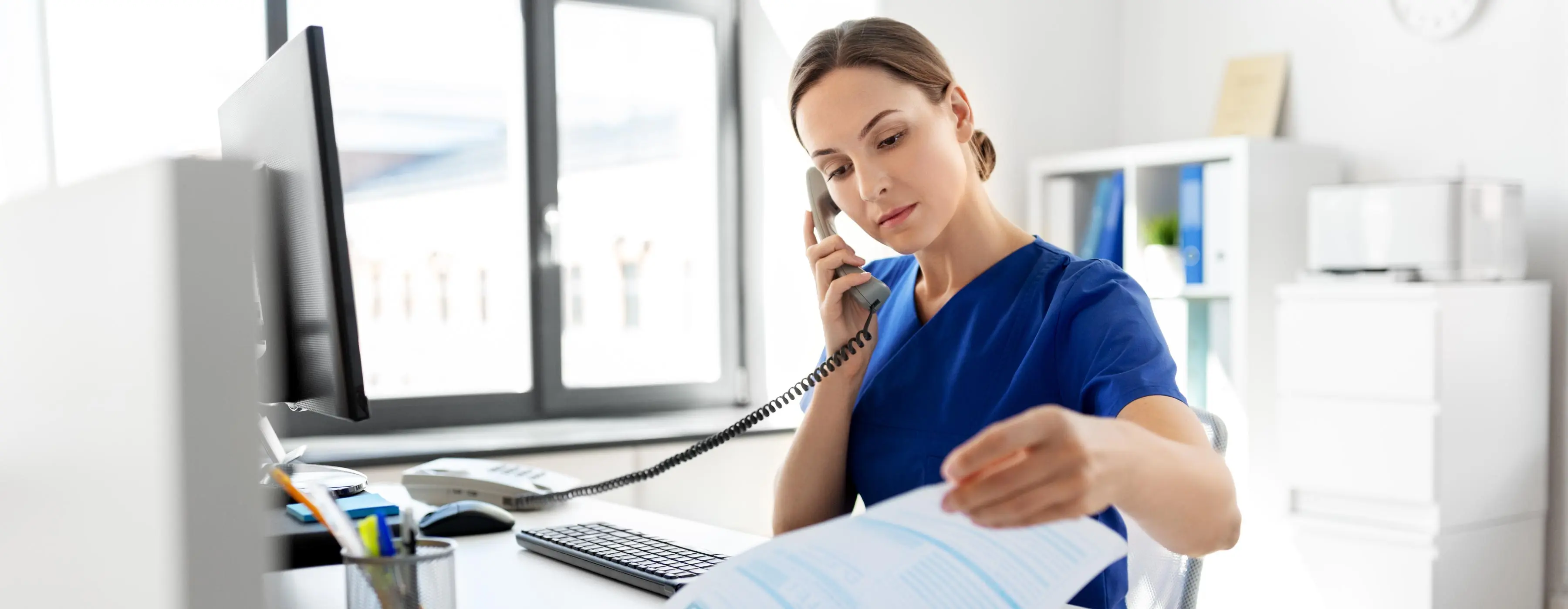 Foto de uma profissional da saúde falando ao telefone e analisando documentos em ambiente de trabalho