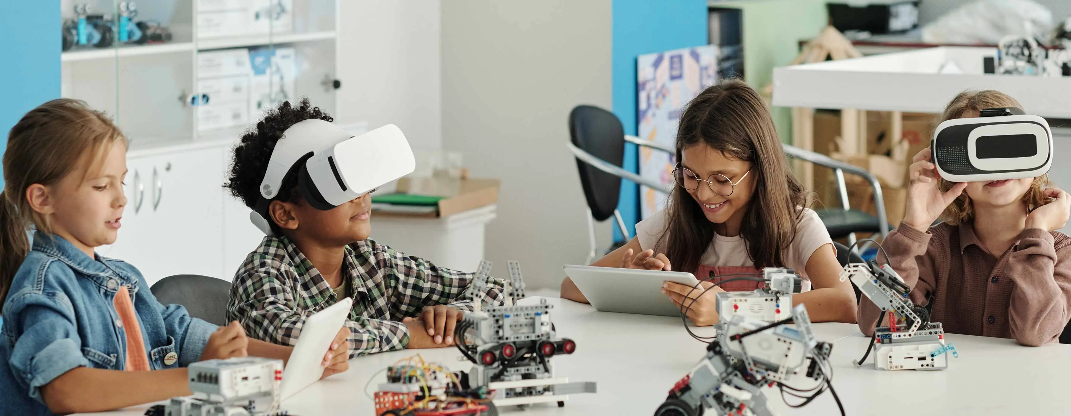 Foto de um grupo de crianças sentadas à mesa, que possui equipamentos robóticos, duas crianças usam óculos de realidade virtual