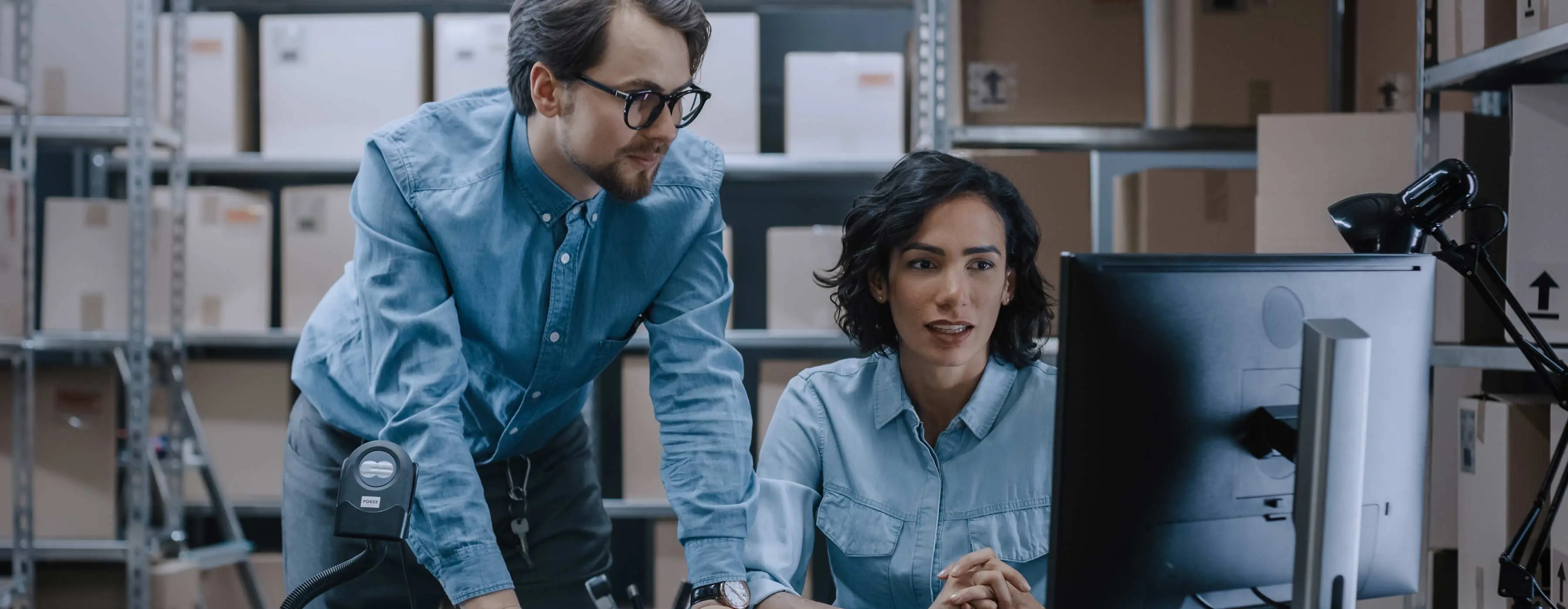 Foto de homem e mulher olhando para computador em ambiente de caixas de papelão em estantes.