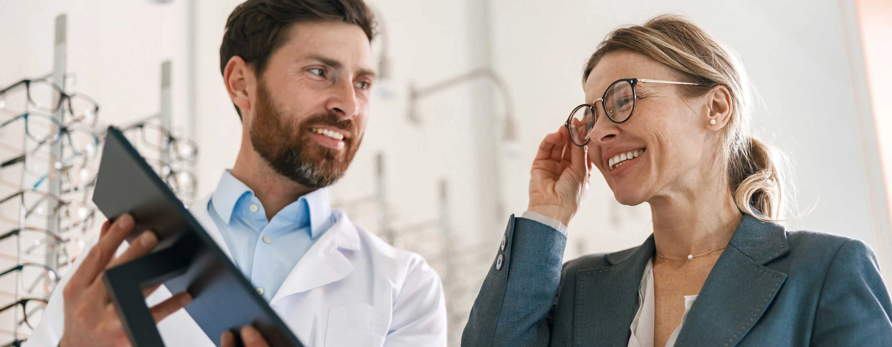 Foto de um homem segurando um espelho para que a mulher ao lado observe um óculos em seu rosto enquanto sorri
