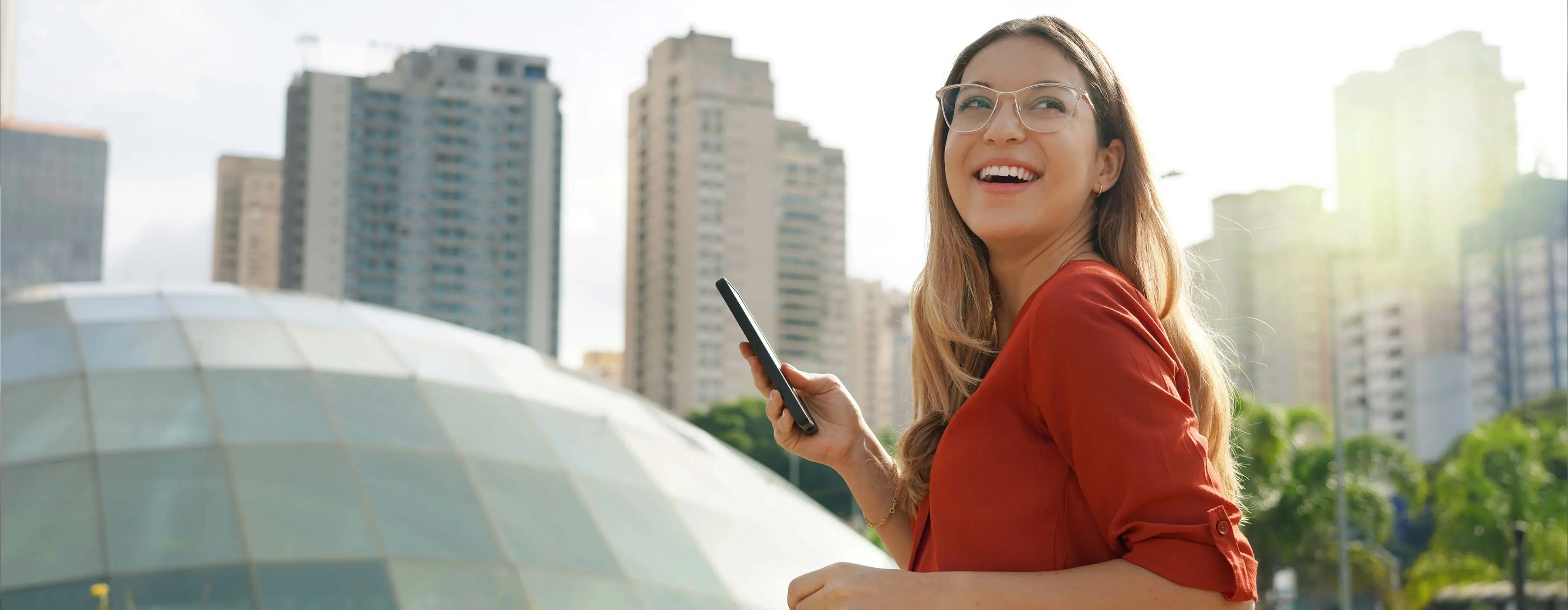 Foto de mulher sorridente usando blusa vermelha e óculos segura celular na mão em ambiente externo com redoma de vidro e prédios atrás junto a árvores.
