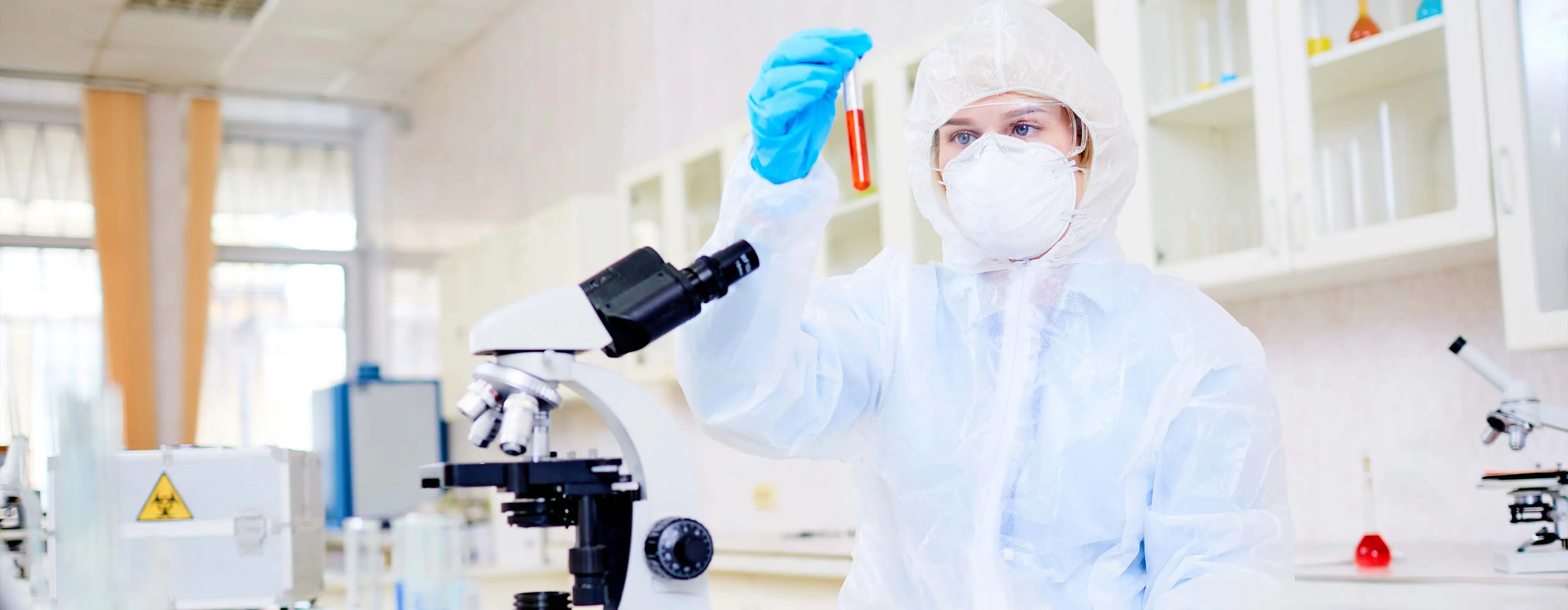 Foto de mulher usando EPIs, máscara e roupa de proteção mexe em tubo de ensaio na frente de uma bancada com um microscópio, tudos de ensaio e computador, em um laboratório
