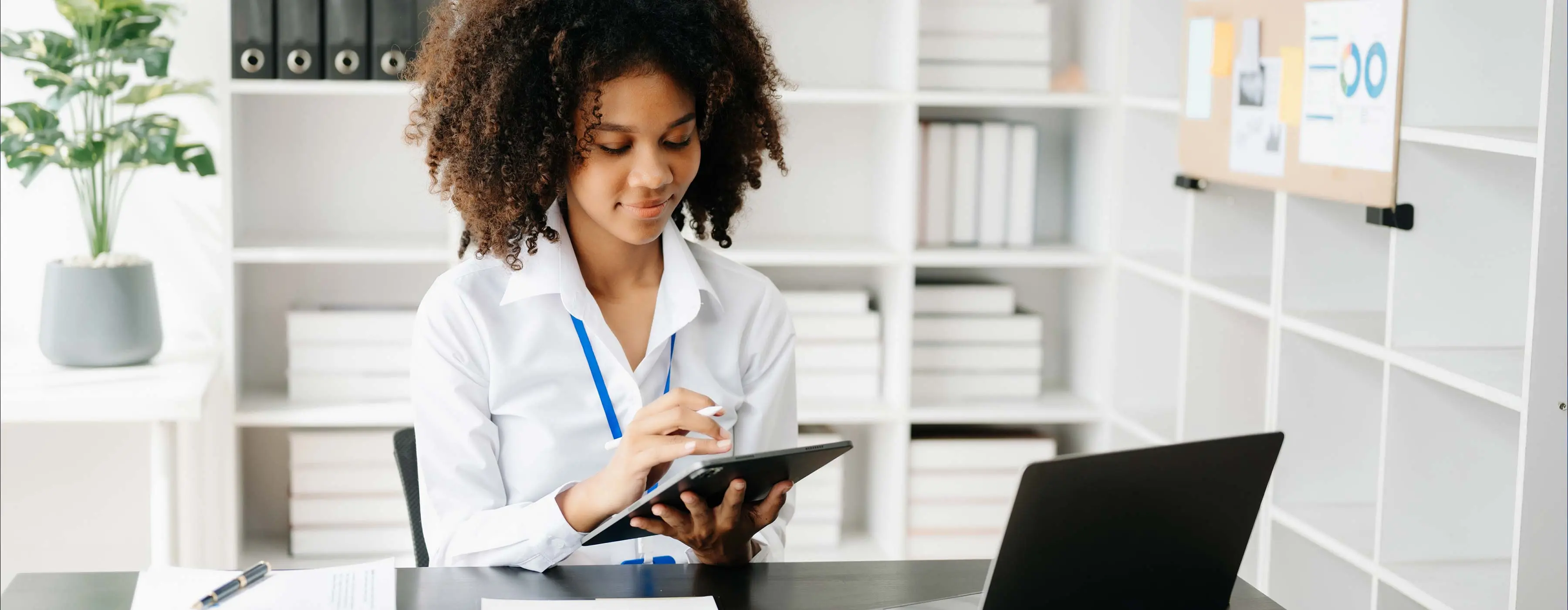 Foto em close de jovem profissional sentada à mesa, mexendo em um tablet em ambiente corporativo