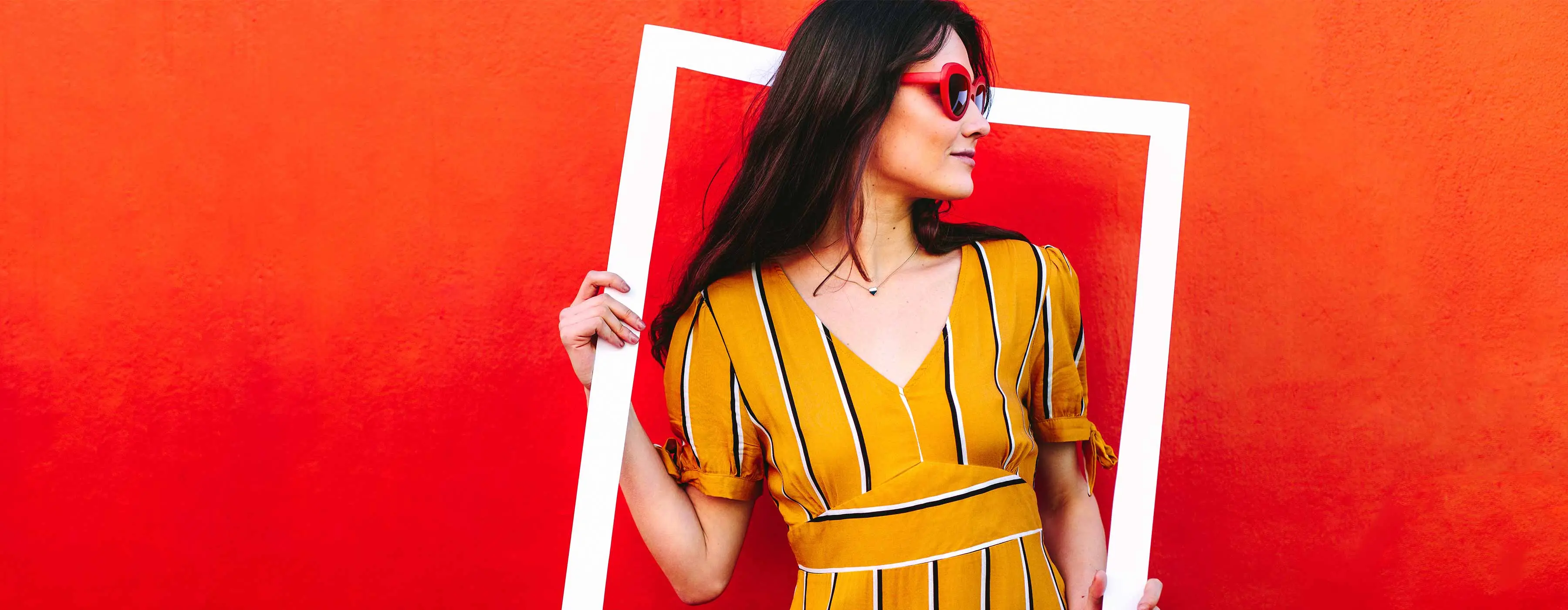 Foto de uma mulher de vestido amarelo e óculos vermelhos segurando um retângulo vazado que enquadra ela em um fundo vermelho