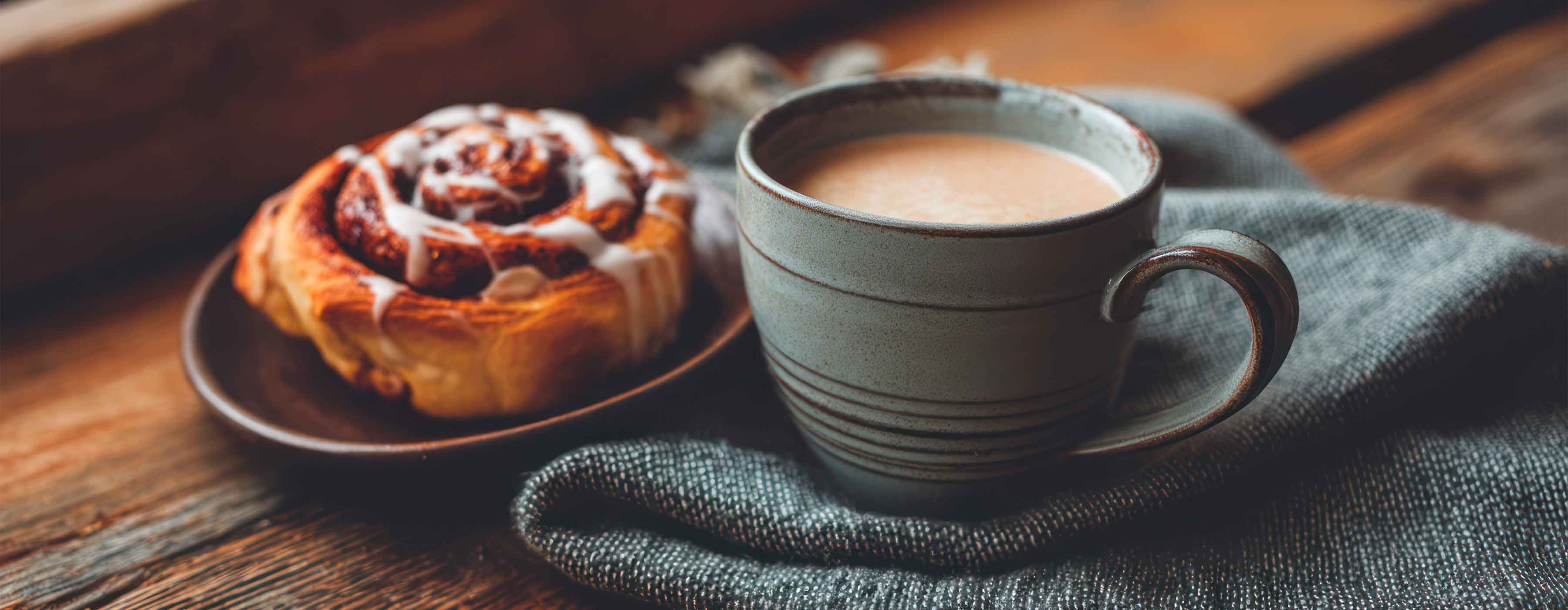 Harmonização com Cafés - foto de uma caneca de cerâmica com bebida quente sobre pano cinza, ao lado de um pão doce, ambos em cima de uma superfície de madeira.