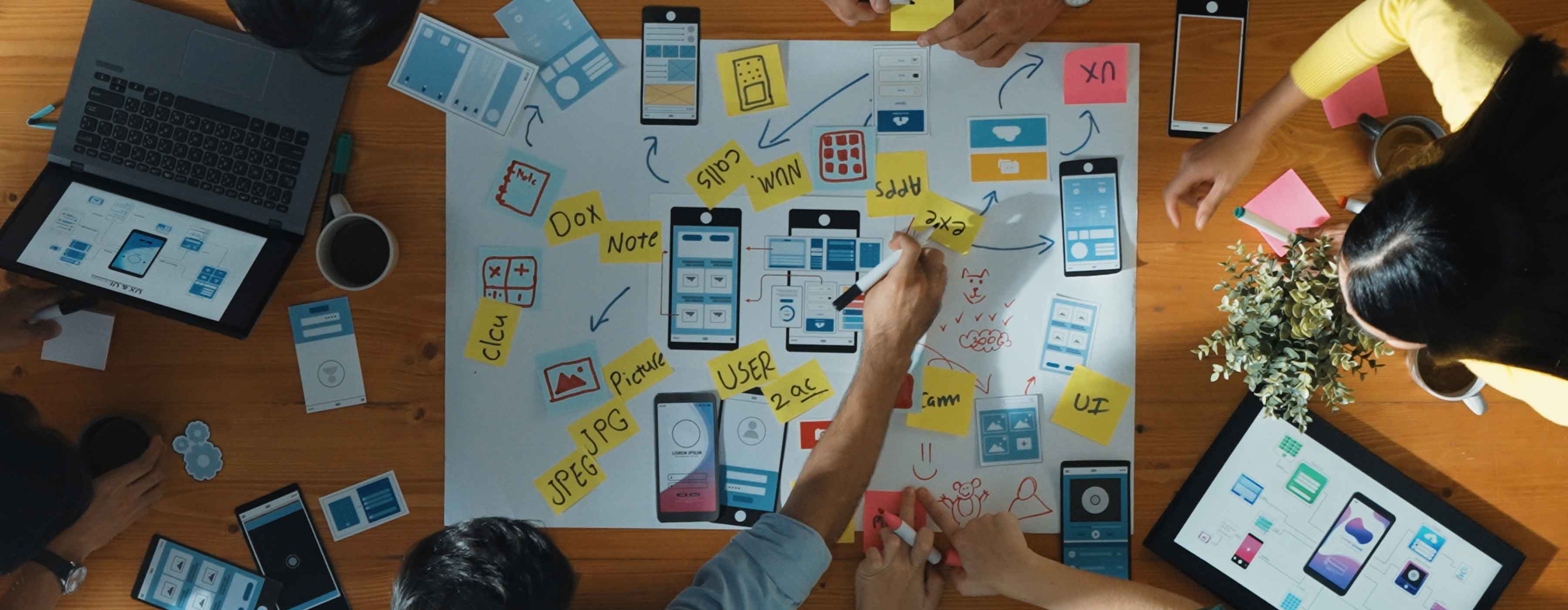 Designer de Experiência do Usuário e Interfaces Digitais - foto de pessoas construindo um protótipo utilizando papéis e post-its ao redor de uma mesa de madeira com notebooks.