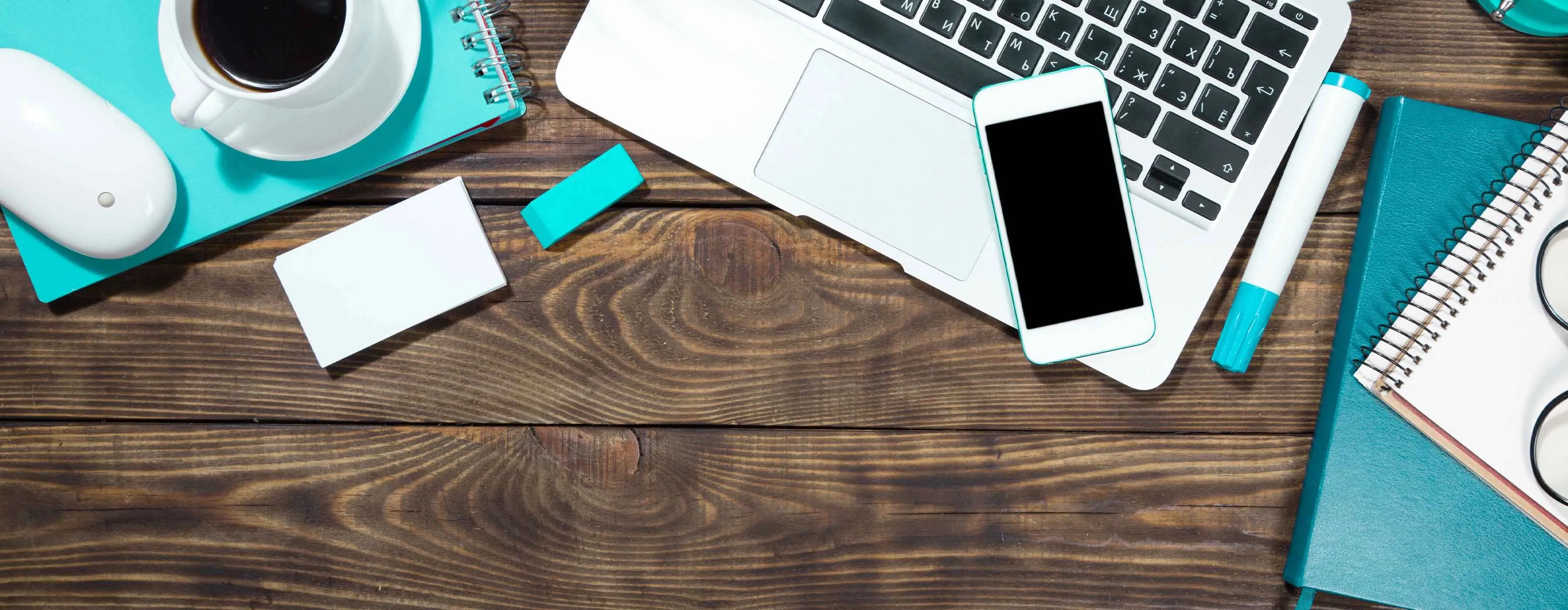 Imagem vista de cima de uma mesa de madeira com notebook, celular, café e outras utensílios.