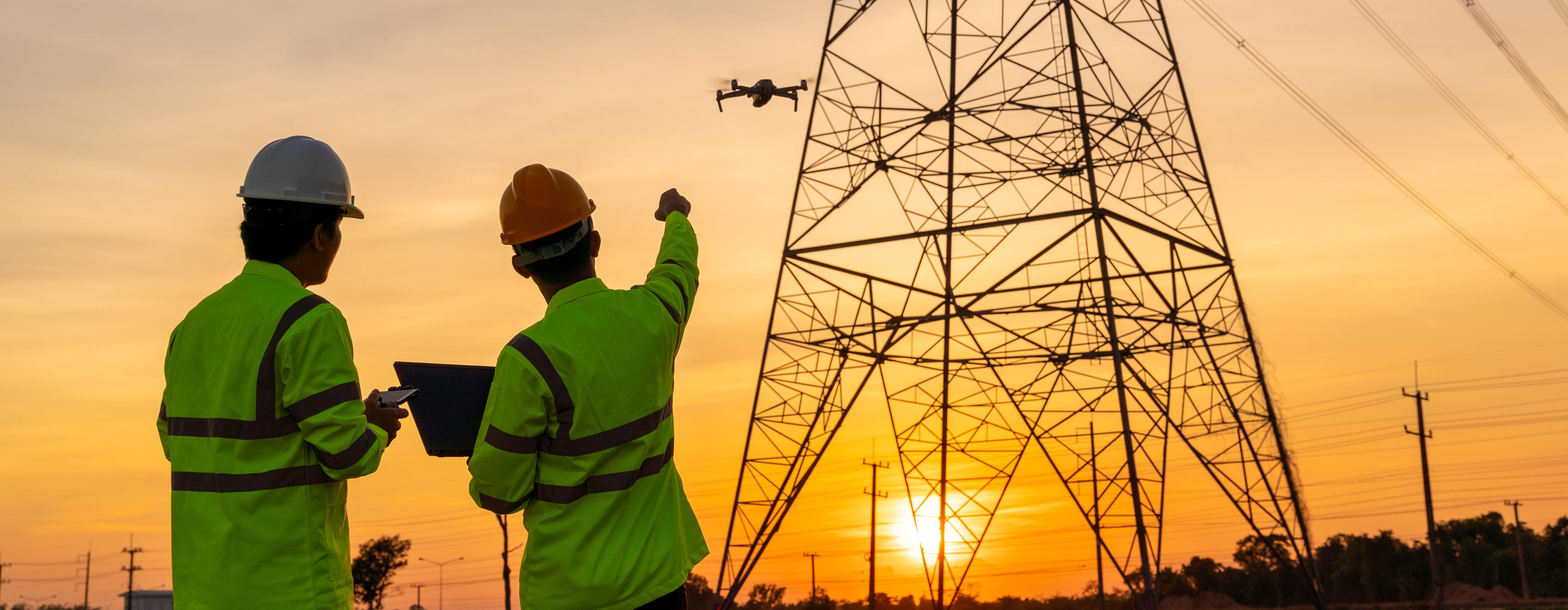 Dois trabalhadores com equipamentos de segurança observam e controlam um drone próximo a uma torre de transmissão de energia, ao pôr do sol.