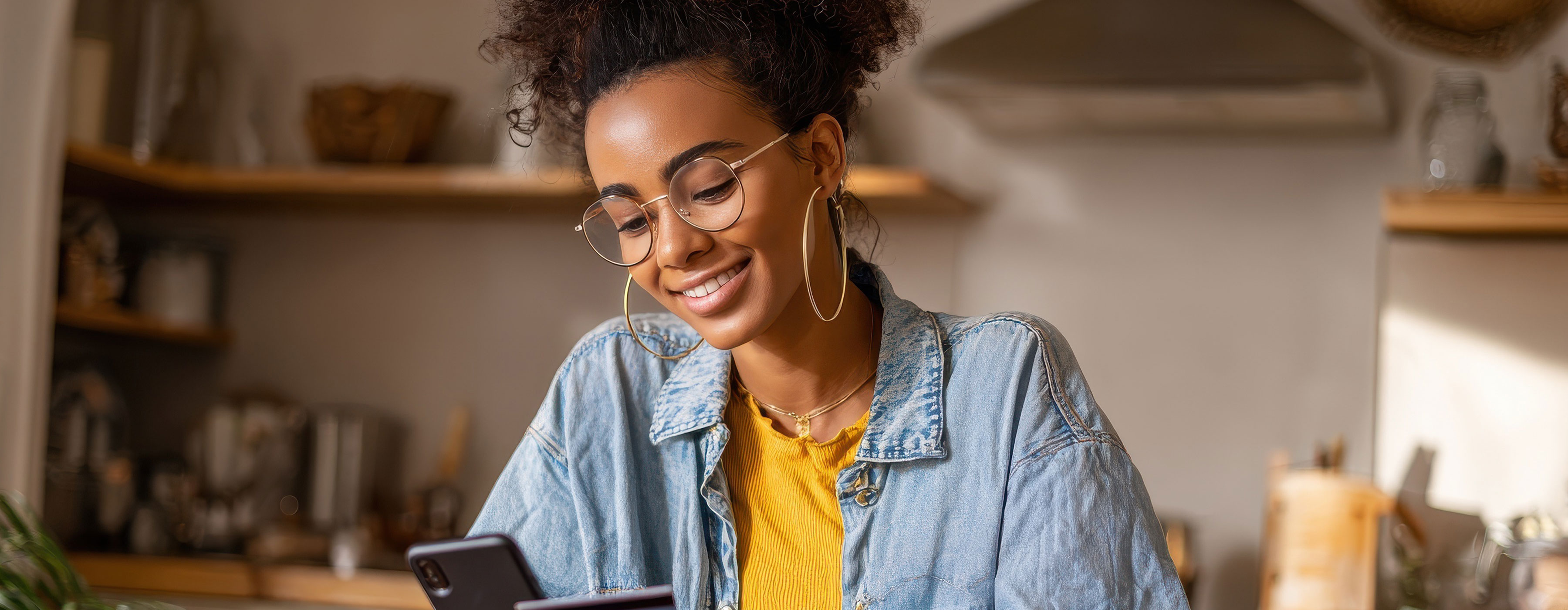 Foto de uma mulher sentada, olhando para a tela do celular e sorrindo. Ao fundo uma imagem desfocada de uma parede com prateleiras e alguns utensílios. 