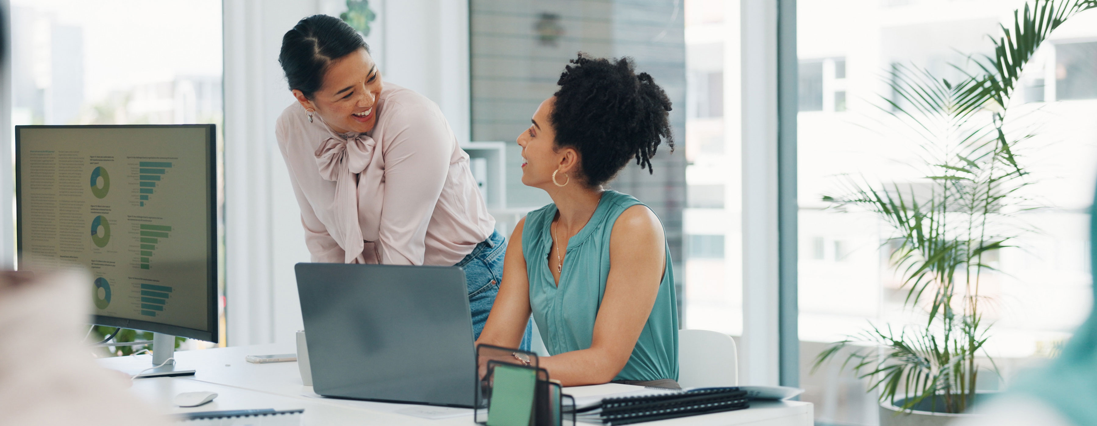 Assistente de E-commerce - foto de uma mulher, sentada à mesa com um laptop, conversa com outra que está em pé ao seu lado.