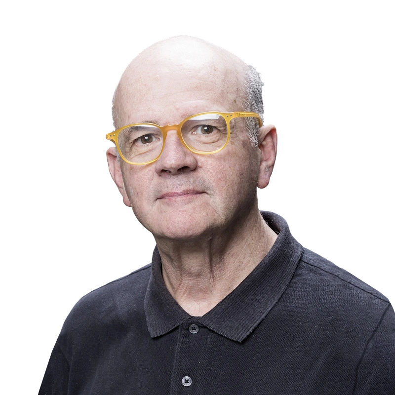 Foto de Guto Lacaz com óculos de grau amarelo e uma camiseta polo preta.