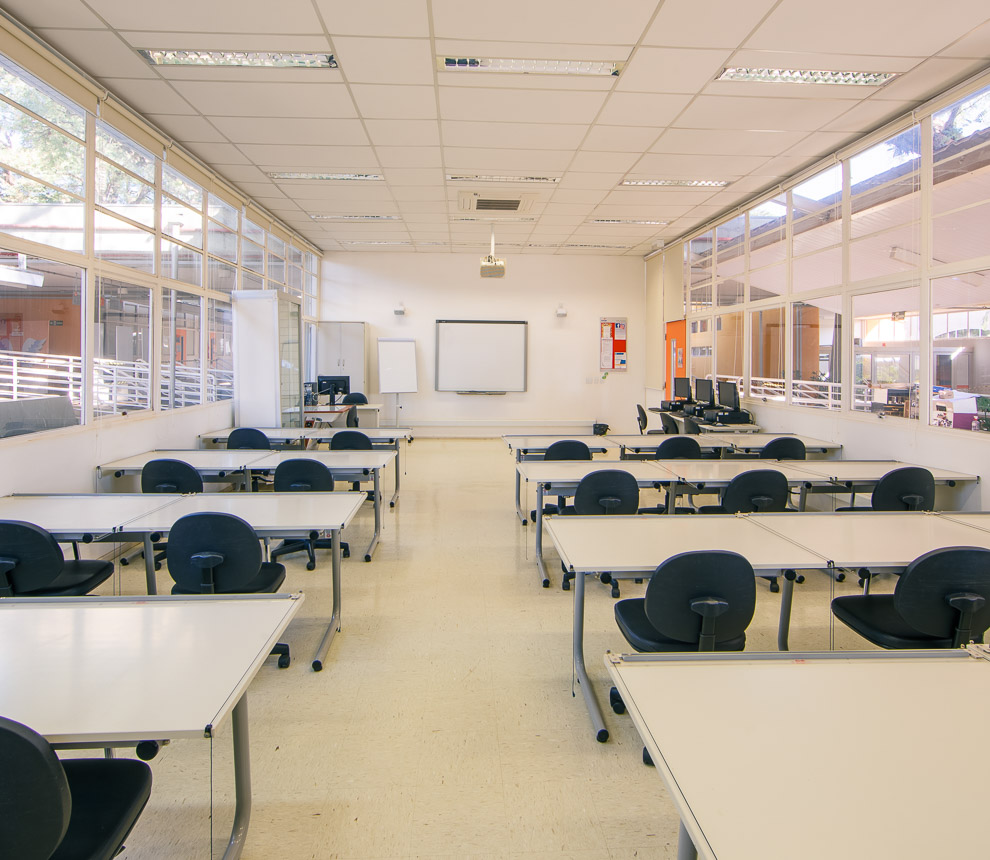 Ateliê de Design possui à esquerda sete mesas-pranchetas de desenho. À esquerda são outras seis mesas-pranchetas para desenho. Ao fundo três cadeiras estão posicionadas em uma mesa convencional em frente à lousa digital. No canto à direita está a porta de entrada e três computadores que podem ser utilizados para pesquisas em aula.