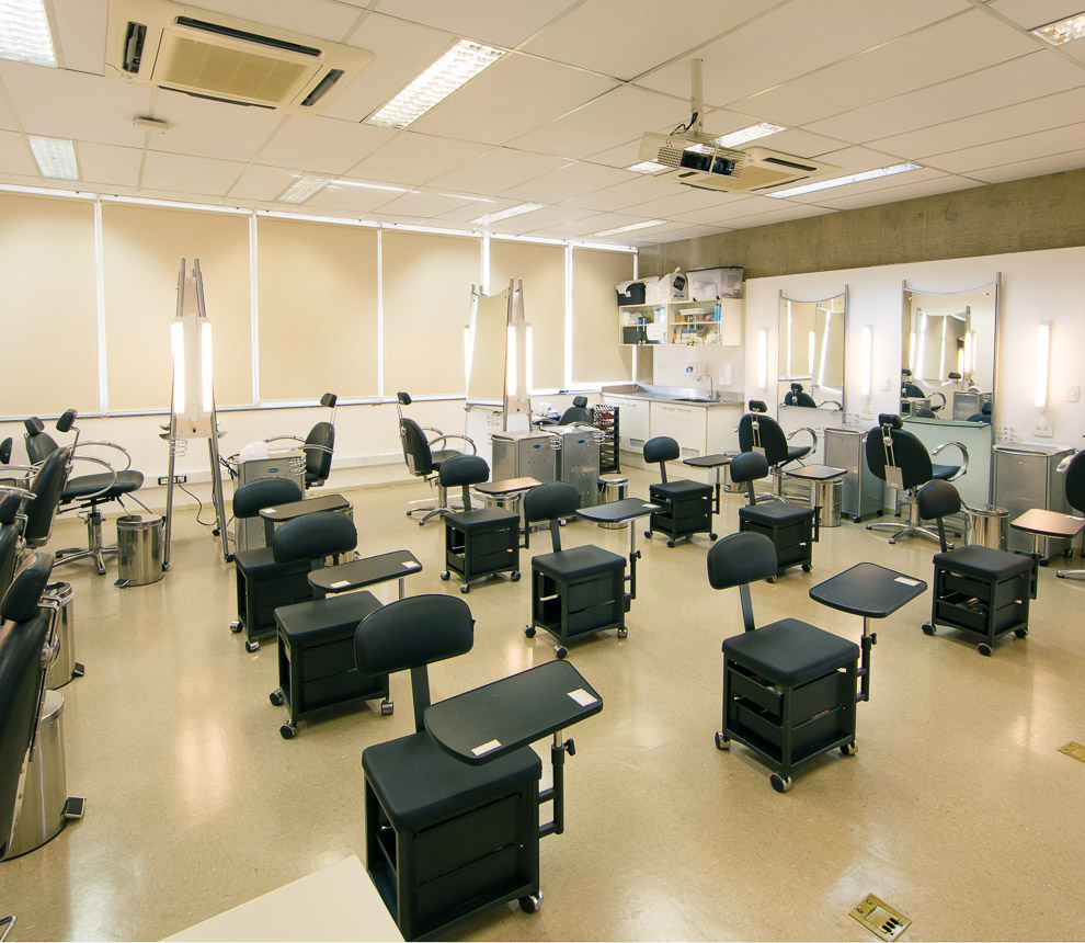 Laboratório de Beleza. Possui sete carteiras convencionais enfileiradas. Ao fundo à direita um armário com rodas e gavetas e uma penteadeira com espelhos e poltrona para procedimentos de maquiagem e sobrancelhas. Ao fundo à esquerda outras três penteadeiras com espelhos e poltronas para procedimentos de maquiagem e sobrancelhas.