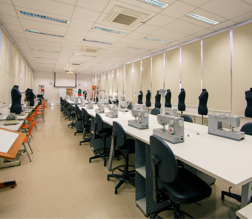 No laboratório de moda possui à direita uma bancada com oito máquinas de costuras posicionadas frente a frente. Em cima da bancada tem também tesouras, carreteis de linhas e revistas de moda. Ao fundo manequins exibem roupas produzidas pelos alunos. O primeiro está no canto à esquerda com um vestido curto em cetim rosa, ao lado, um vestido curto verde militar e por último um vestido preto com cortes assimétricos.