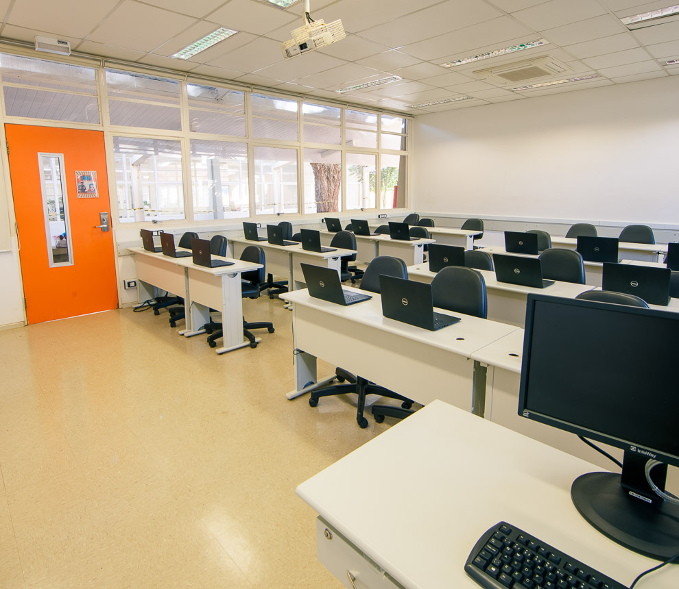 Laboratório Móvel com quatro bancadas posicionadas à direita. Em cima de cada bancada tem 2 notebooks para uso de alunos. À esquerda outras quatro bancadas com três notebooks em cima de cada uma. Em frente de cada notebook no chão estão posicionadas cadeiras com rodinhas e almofadadas.