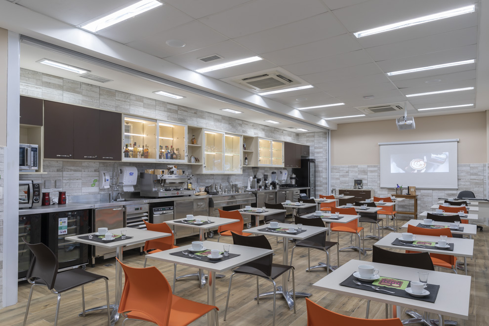 Imagem do Laboratório de Sala e Bar, com equipamentos para fazer café, geladeira para cervejas, prateleira de bebidas, taças, copos, xícaras, além de mesas e cadeiras enfileiradas. Ao fundo tem o retroprojetor e a mesa para uso do docente