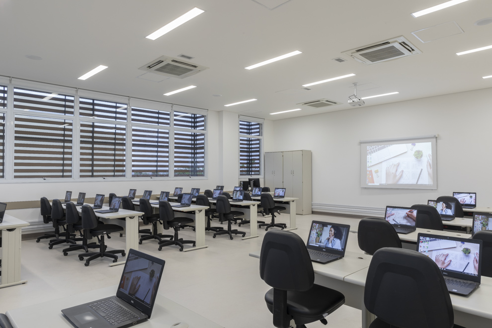 Rodeada de janelas, a sala multimídia do Senac Salto tem diversas mesas e notebooks. Ao fundo, o projetor na parede e a mesa do docente.