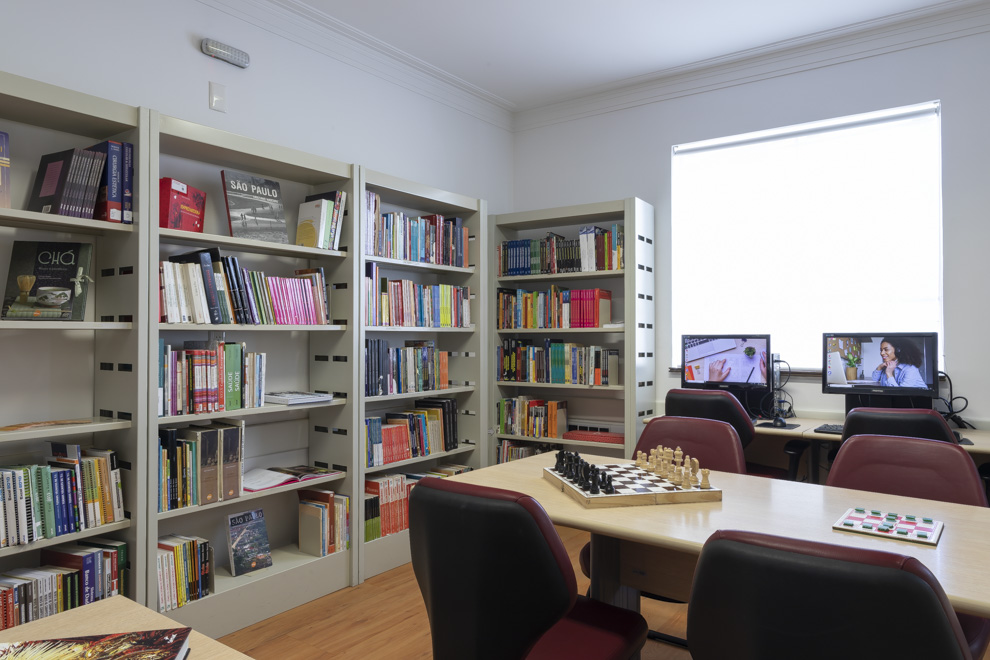 Foto com mesa com jogos em cima, cadeiras, estantes cheias de livros e computadores para acesso à internet