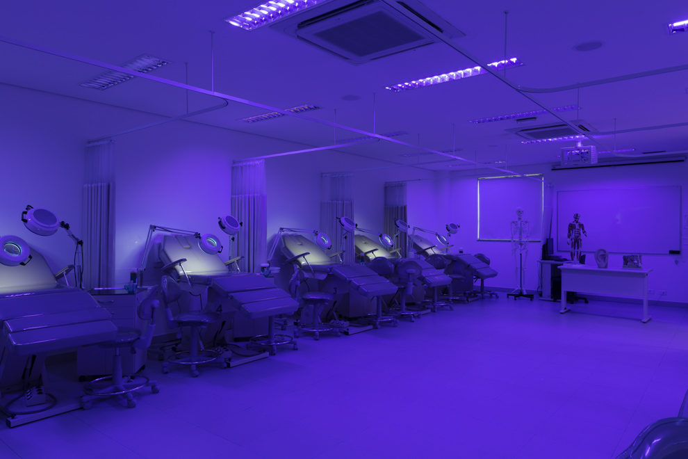 Laboratório com luz azul, com diversas macas de atendimento enfileiradas e equipamentos para realização de limpeza de pele, no fundo há esqueletos para estudo da anatomia humana e um quadro onde está desenhada uma estrutura celular