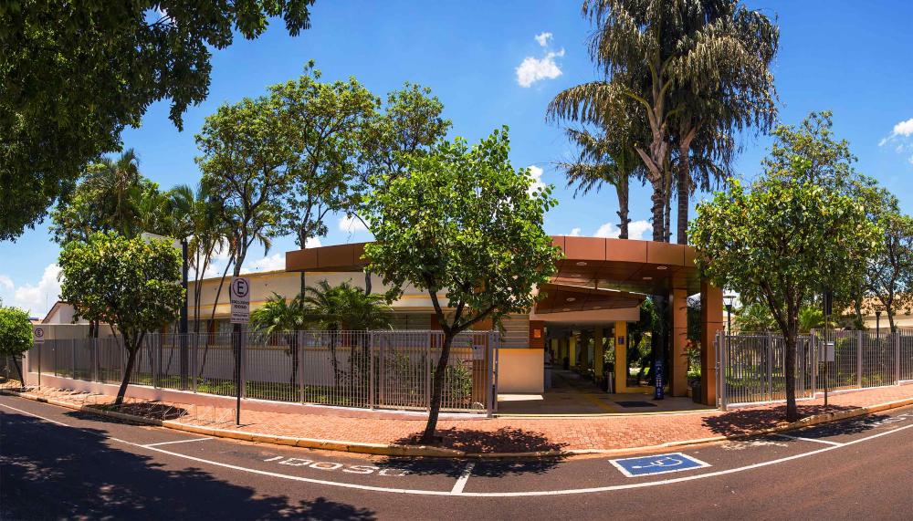 A foto da fachada da unidade aconteceu ao entardecer e as luzes estão acesas. À esquerda há uma árvore na calçada e um pequeno jardim com palmeiras e plantas em vasos. À direita há uma árvore na calçada e um jardim extenso com palmeiras pequenas e grandes.