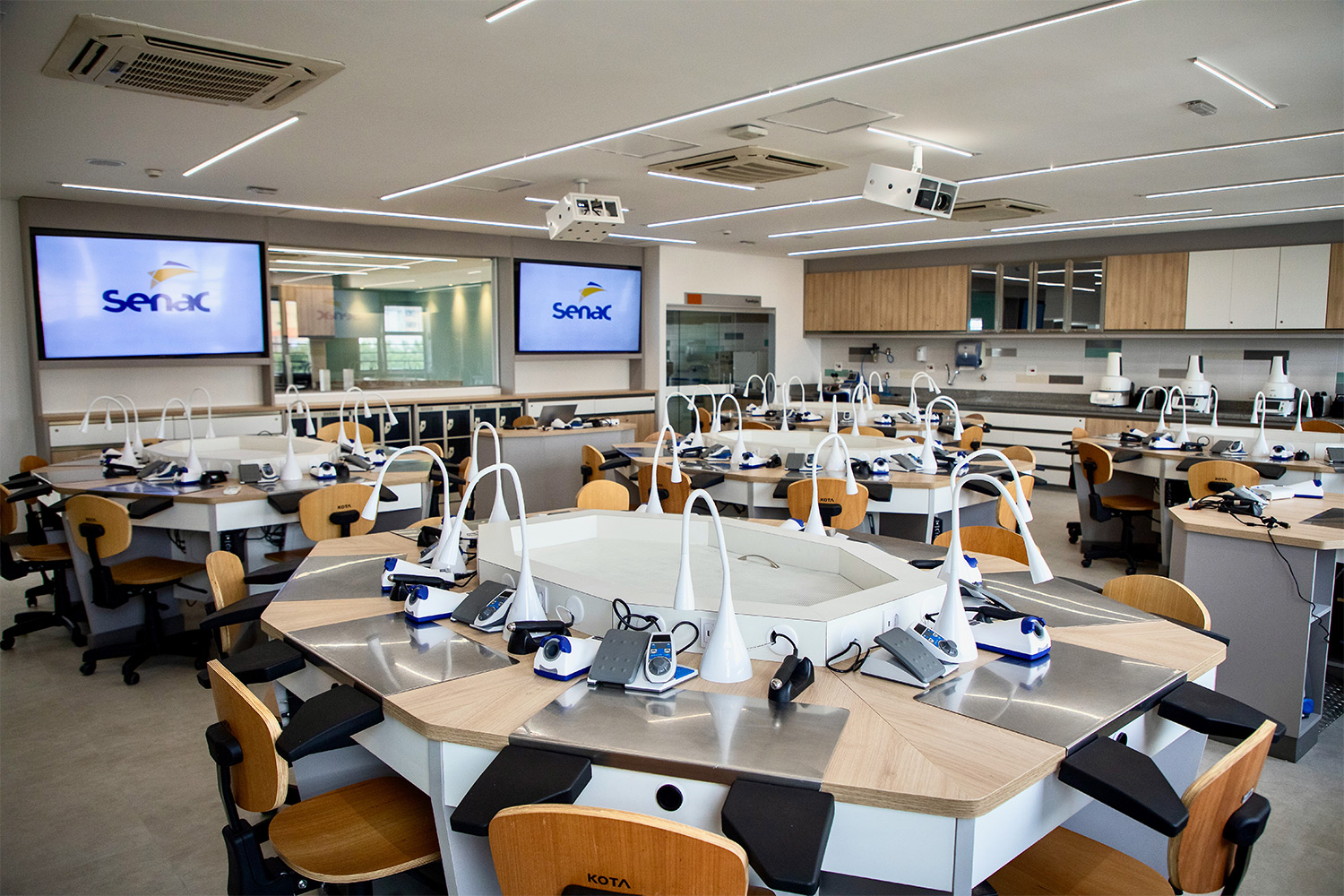 Foto geral do laboratório de Prótese Dentária, com bancadas, cadeiras, equipamentos e duas televisões.