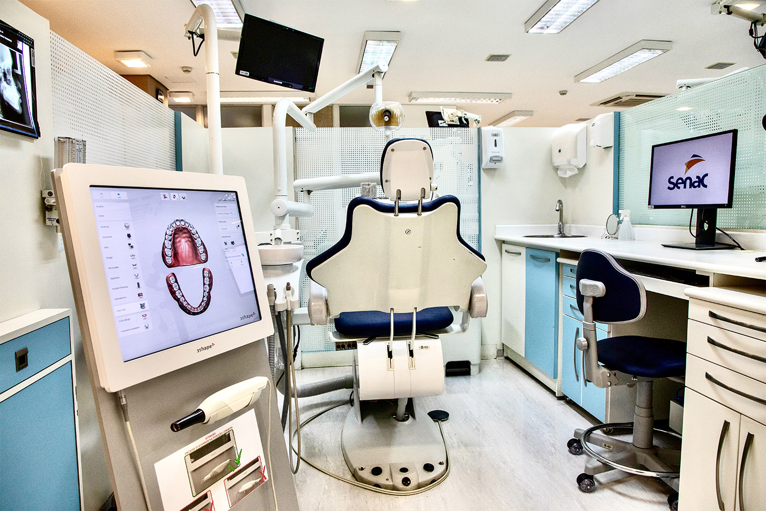 Foto de destaque a monitores ligados ao scanner intraoral da clínica. A imagem também mostra cadeira e ferramentas utilizados em atendimentos odontológicos.