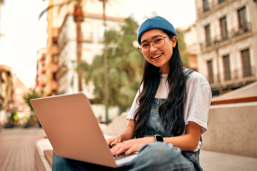 Imagem: uma estudante sentada na escadaria de uma praça pública sorri enquanto estuda com um laptop apoiado nos joelhos.