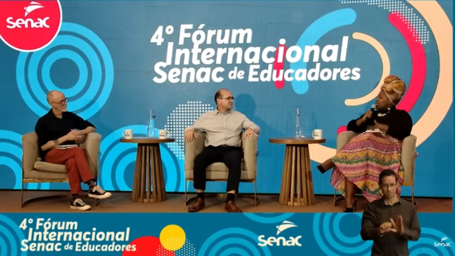 Três pessoas sentadas em poltrona posicionadas lado a lado no palco. Uma pessoa fala ao microfone e as outras duas olham para ela.