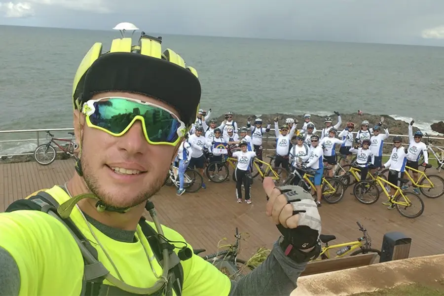 Fagner está tirando uma selfie durante um passeio de bicicleta à beira-mar. Ele usa capacete preto e amarelo, óculos esportivos com lentes espelhadas e camiseta amarelo-neon, apontando para um grupo de cerca de 20 ciclistas ao fundo. Os participantes estão posicionados em um deck de madeira próximo a uma área rochosa, com bicicletas amarelas e camisas brancas. Ao fundo, aparece o oceano sob céu nublado.