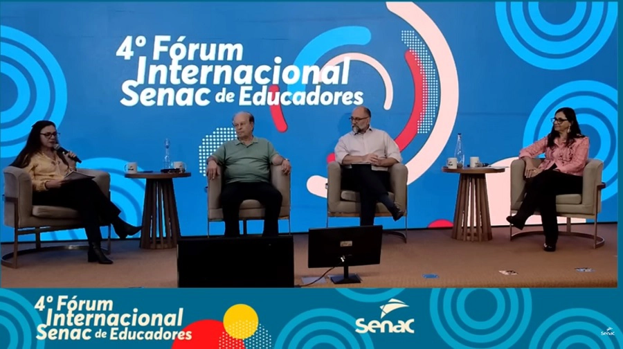 Foto: imagem mostra quatro pessoas sentadas em cadeiras em um palco durante o 4º Fórum Internacional Senac de Educadores. Ao fundo, há um painel azul com o logotipo do Senac no canto superior esquerdo e o título do evento centralizado.