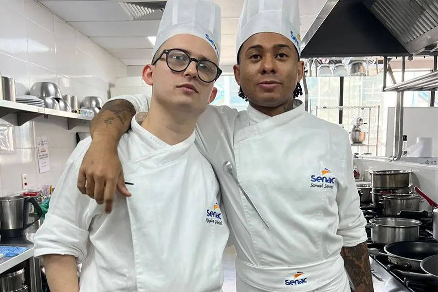 Nikolas (esq) e Samuel estão abraçados vestindo dólmãs brancas no laboratório de Gastronomia do Centro Univertsitário Senac.