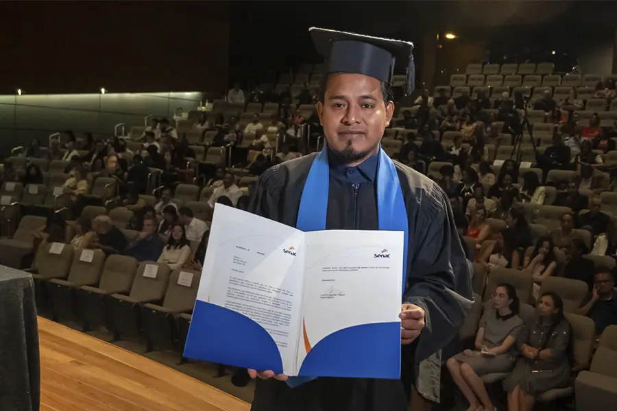 Ramon Ferreira está no auditório do Centro Universitário Santo Amaro segurando a documentação referente à sua formatura. Ele usa beca preta e capelo.