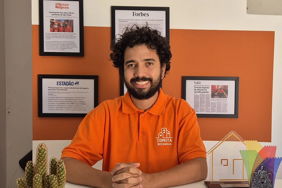 Imagem de Sidney Bruno. Ele está sentado sorrindo, e está usando uma camisa polo laranja com uma inscrição "Comuta Reformas". Ao fundo, uma parece traz quadros com notícias publicadas sobre o projeto.