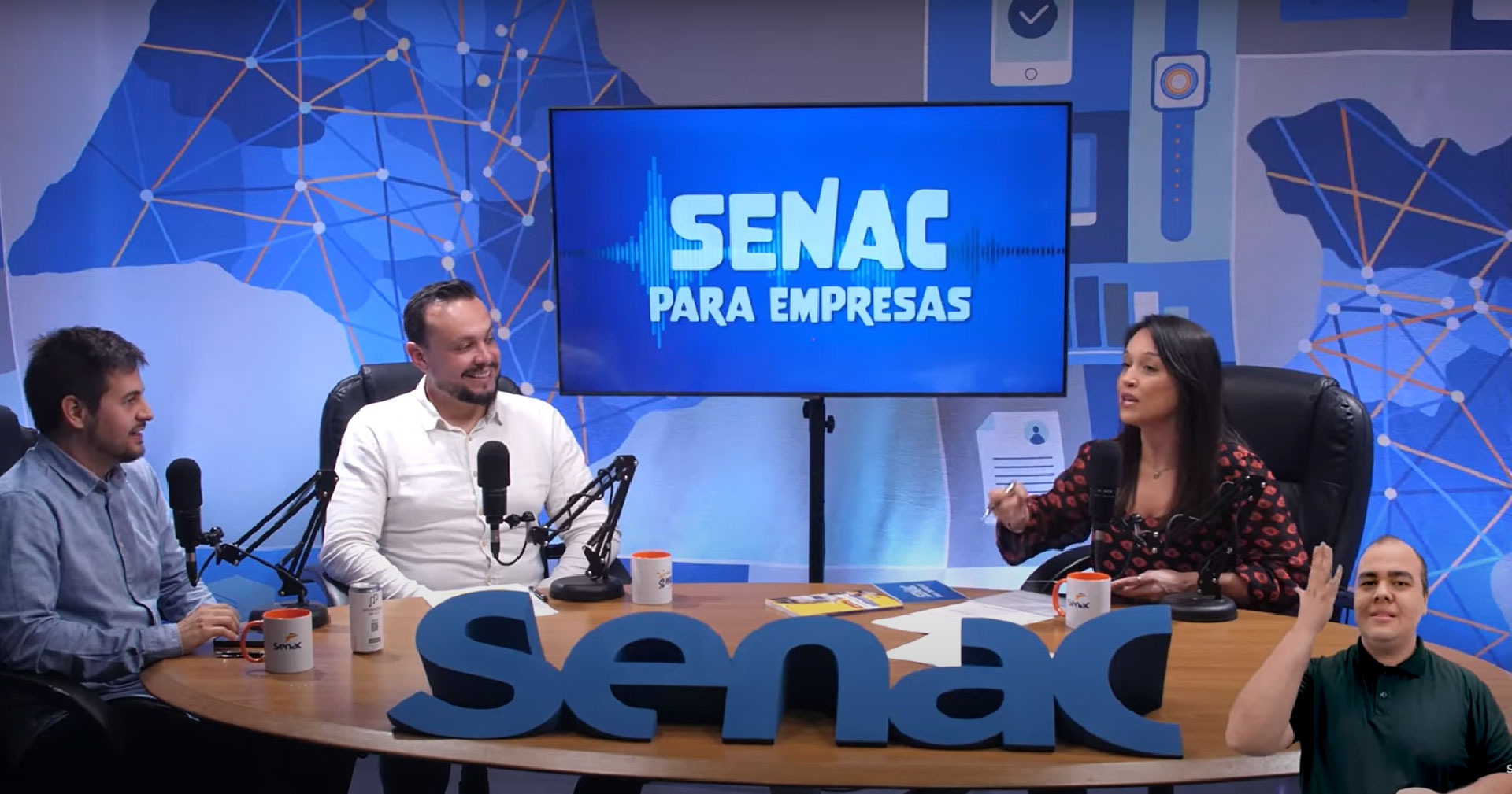 Gravação de podcast com três pessoas sentadas ao redor de uma mesa, cada uma com microfone. Ao fundo, painel colorido e tela exibindo o nome: Senac para empresas. Na mesa, canecas com o logo do Senac. Ambiente profissional voltado à discussão de temas educacionais ou corporativos.