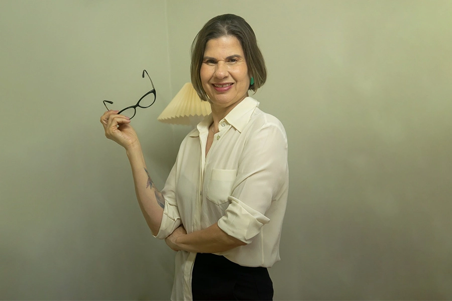 Imagem de Cleusa sorrindo para a câmera. Ela está em pé, com um braço cruzado e outro segurando um óculos.