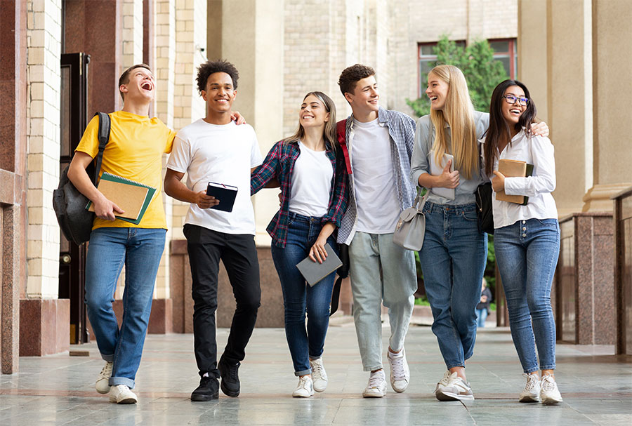 Imagem:  grupo sorridente de jovens estudantes de diferentes raças e gêneros abraçados lado a lado.