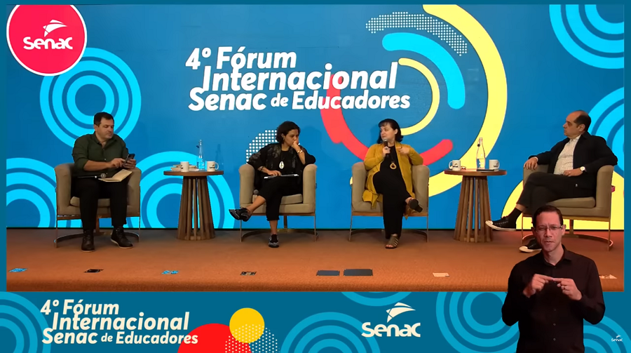 Imagem: quatro pessoas sentadas em cadeiras em um palco durante o 4º Fórum Internacional Senac de Educadores. Ao fundo, há um painel azul com o logotipo do Senac no canto superior esquerdo e o título do evento centralizado.