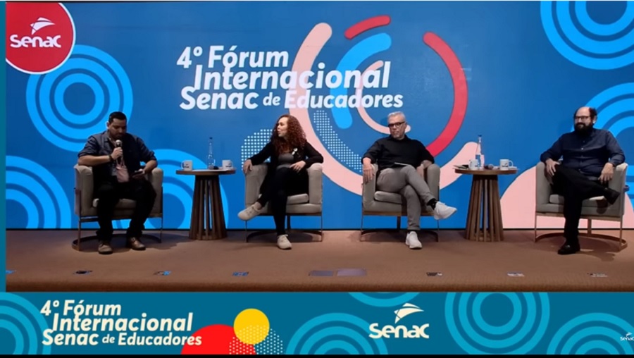 Foto: imagem mostra quatro pessoas sentadas em cadeiras em um palco durante o 4º Fórum Internacional Senac de Educadores. Ao fundo, há um painel azul com o logotipo do Senac no canto superior esquerdo e o título do evento centralizado.
