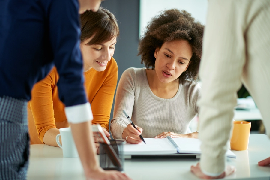 Imagem: três mulheres de um grupo de trabalho escutam uma colega do grupo enquanto ela explica a ideia do projeto e aponta para um caderno com anotações.